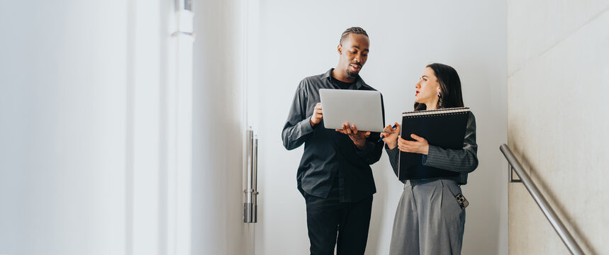 Two business people collaborating in a modern office environment, using a laptop and notepad for planning. Promoting teamwork and productivity in a diverse workplace setting.