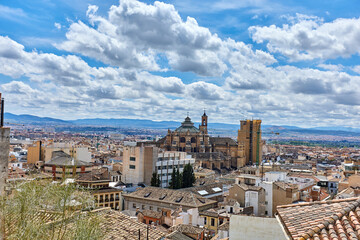 Obraz premium Granada cityscape with tiled rooftops, clustered buildings, and a large cathedral with domes and towers under a sky filled with scattered clouds and distant hills