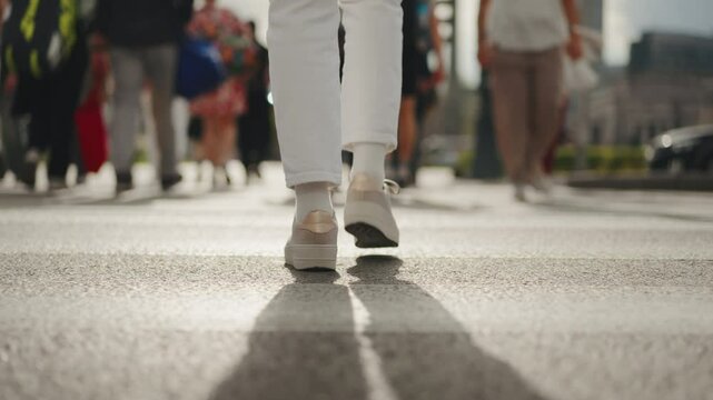 Back view following shot female feet steps walking pedestrian crossing street, sunny day, young woman wearing white pants and sneakers goes crosswalk. Low angle people legs stepping zebra asphalt