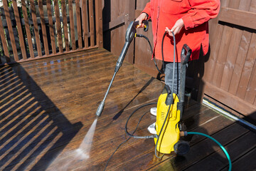 Person Using High-Pressure Washer to Clean Wooden Decking Boards in front of Brown Garden Fence