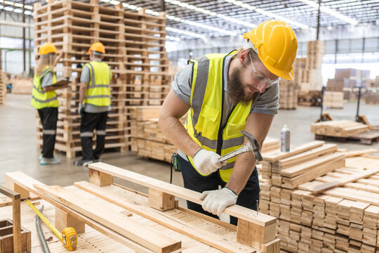 Male workers carpenter wearing safety uniform and yellow hard hat working using hammer nail to on wood at workshop. Man worker wood surrounded piles of wooden pallets in warehouse industry.