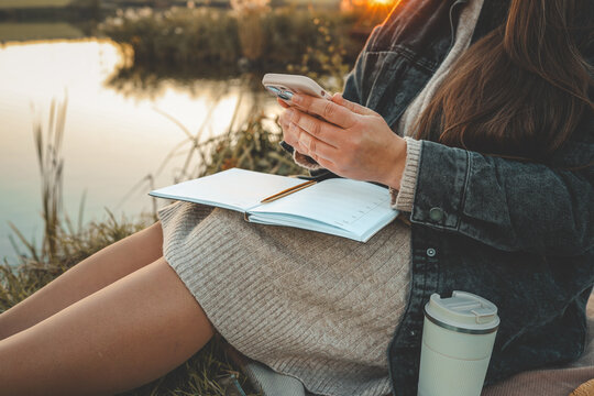 Woman holding smartphone while sitting outdoors with planner and pencil on lap. Cozy autumn atmosphere near lake, with coffee cup resting nearby