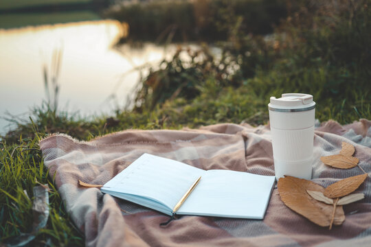 Open planner and reusable coffee cup on a plaid blanket near a peaceful lake at sunset, with fallen leaves adding an autumn touch