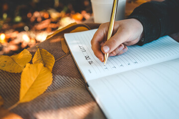 Hand writing in a planner with a gold pen, surrounded by yellow autumn leaves, a reusable cup, and cozy textures, creating a warm seasonal to-do list scene