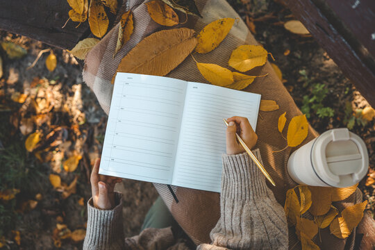 Hand writing in a planner with a gold pen, surrounded by yellow autumn leaves, a reusable cup, and cozy textures, creating a warm seasonal to-do list scene