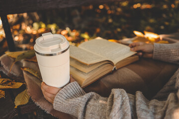 Person in cozy sweater holding reusable cup while reading a book outdoors in autumn park, surrounded by warm tones, fallen leaves, and soft sunlight