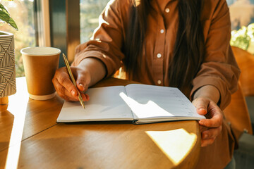 Woman writing in open planner at sunny café table with takeaway coffee cup and houseplants,...