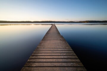 Fototapeta premium A tranquil wooden pier extends across a calm lake, reflecting the clear blue sky and surrounding nature at dawn or dusk.