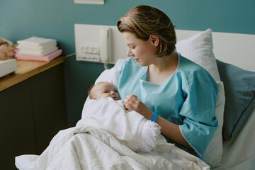 New mother holding her newborn baby while lying in hospital bed, wrapped in blankets and next to...