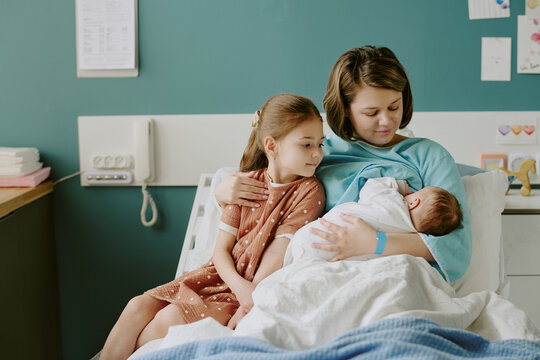 Young girl gently embracing sibling while mother cuddles newborn on hospital bed in welcoming environment with natural light illuminating room medical setting