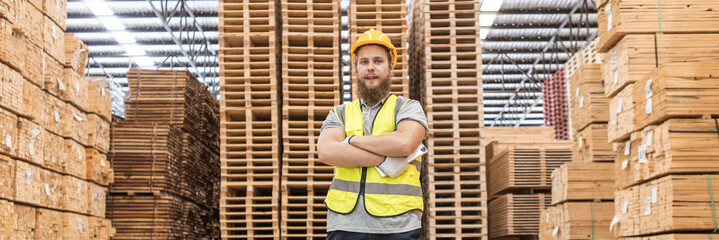 Portrait man worker wearing safety uniform and yellow hard hat standing crossed arms looking at camera smile surrounded piles of pallets background in wooden warehouse.