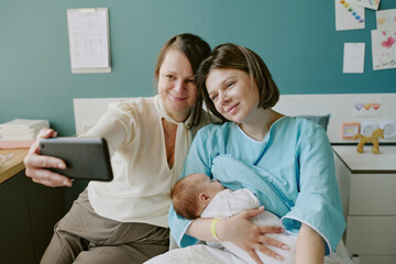 Portrait of grandma, mother, and newborn baby bonding while capturing selfie. Smiling grandma holding phone, seated next to daughter in hospital with infant on mother's lap
