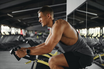 Side view of handsome guy exercising on stationary cycle in modern gym while holding handle bar...