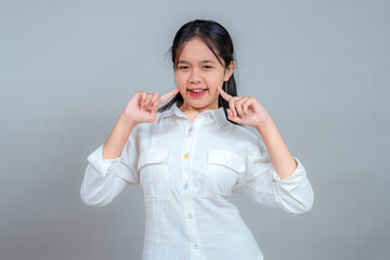 Smiling Asian young woman pressing her index fingers to her cheeks while winking playfully, showing...
