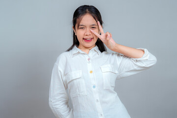 Playful young Asian woman winking with a cheerful smile while making a peace sign near her eye,...