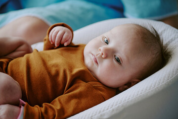 Portrait of baby wearing brown shirt and resting on bed while looking up with a relaxed expression,...
