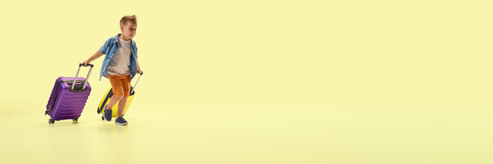 Banner. Boy confidently pulling yellow and purple luggage looking forward to vacation fun against yellow background with negative space. Concept of childhood, travel, summer, lifestyle, joy.