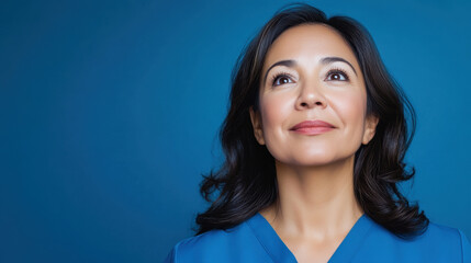 Confident middle aged woman with dark hair wearing blue top looks upward with hopeful expression against solid blue background