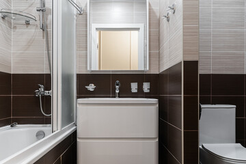 small bathroom with white bathtub, white sink, square mirror, cabinet under sink, chrome faucet. brown and beige ceramic tiles on walls. Bathroom design in flat, hotel or apartment.