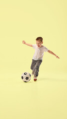 Athletic little boy, playful champion kicks soccer ball forward in motion while running across pastel yellow studio background. Concept of vacation, childhood, summer, sport, wellness, energy.
