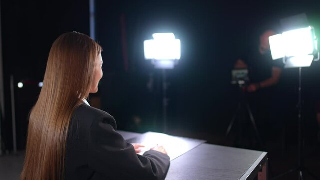 Positive brunette woman with long straight hair sits at the desk half-back to camera. Lights of soffits illuminate the blogger recording a video. Male photographer at backdrop.