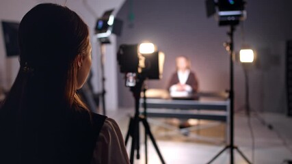 Rear view of a long-haired girl standing at backstage in the photo studio. Lady looks at camera display filming a lady sitting at desk. Blurred backdrop. - Powered by Adobe