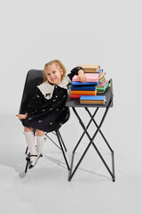 Girl in black dress leans cheerfully on stack of colorful books, smiling confidently at camera. Library. Concept of reading joy, school life, library knowledge, new ideas, and playful education.