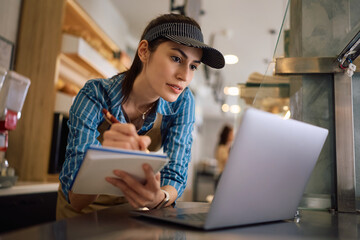 Female bakery shop owner taking notes while working on laptop.