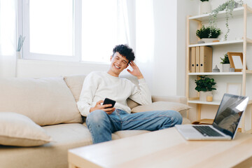 Fototapeta premium young man relaxing at home, sitting on a sofa with a smartphone and laptop, bright cozy interior, natural light through windows, modern decor, casual outfit and cheerful expression