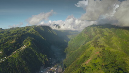 Obraz premium Stunning aerial view of lush green valley with small village nestled amidst towering mountains, with clouds over hill peaks, creating a mesmerizing landscape in the heart of Tahiti, French Polynesia
