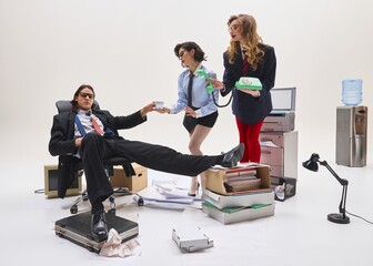 Office man being served coffee by two female colleagues while lounging at desk in chaotic setting....