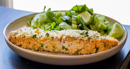 Close-up of a baked breaded salmon fillet served with a simple green salad in a dish. The salmon has a golden, crispy crust and is paired with fresh greens