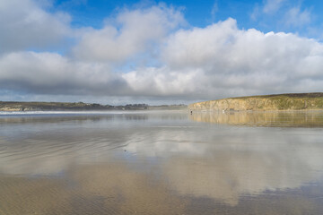 Reflets des nuages blancs sur le sable mouillé à marée basse, avec en arrière-plan les falaises de la presqu’île de Crozon sous un ciel bleu lumineux.