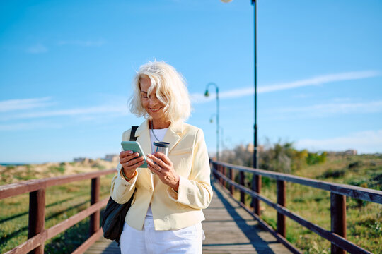 Senior woman enjoying a walk by the seaside while checking her phone - Powered by Adobe