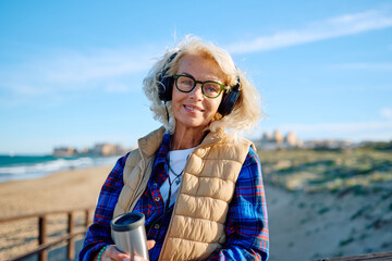 Older woman enjoying morning beach walk with coffee and headphones
