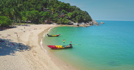 Sandy beach with colorful traditional thai boats, turquoise water, tropical greenery, and relaxing people under a sunny sky, concept of paradise. Thailand, Koh Phangan Island, Haad Thong Reng Beach