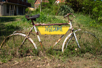 An old, white-painted bike with a yellow "WELCOME" panel is set in a green meadow near traditional homes, creating a cozy, countryside ambiance ideal for rural tourism and homestay branding.