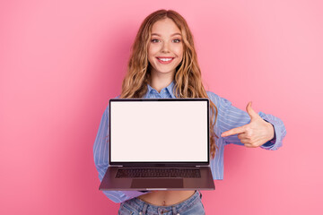 Naklejka premium Smiling young woman in a striped blouse holding and pointing at a laptop with a blank screen on a vibrant pink background