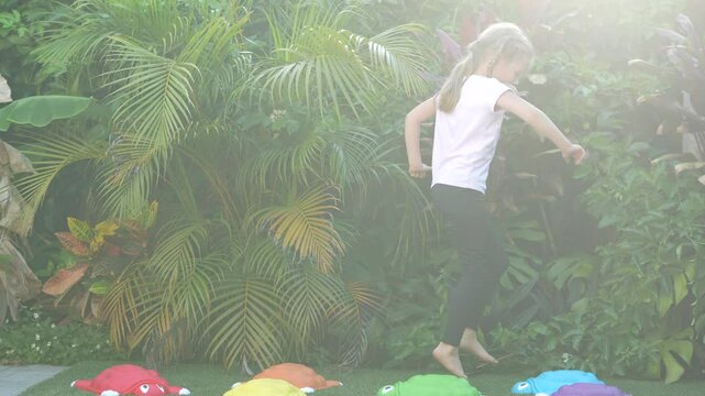 Little girl running and playing outside on artificial grass with colorful turtle stepping stones, tropical plants background, barefoot childhood activity, outdoor fun and movement, candid moment
