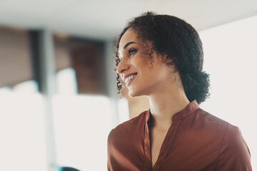 Confident young businesswoman in casual shirt smiling brightly in an office space with natural...
