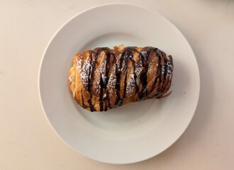 Overhead view of a chocolate-drizzled pastry topped with powdered sugar, perfectly centered on a white plate against a clean, minimalist background.