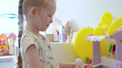 Cute blonde girl with braided hair playing with a colorful ice cream shop toy in her bright, tidy, modern bedroom, surrounded by balloons and other toys