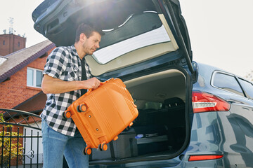 Man loading orange suitcase into car trunk before journey. Packing luggage for travel