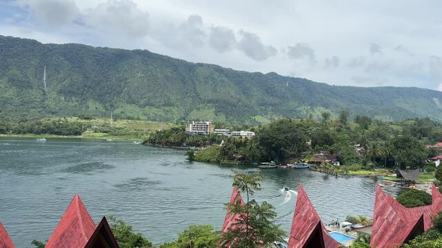 Scenic footage of Lake Toba in North Sumatra, Indonesia, featuring calm waters, traditional Batak rooftops, lush green hills, and a distant waterfall under partly cloudy skies.
