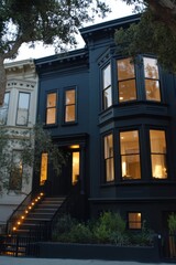 An elegant black Victorian house stands gracefully on a tranquil street, its warm yellow lights glowing invitingly from the windows during the enchanting twilight hours