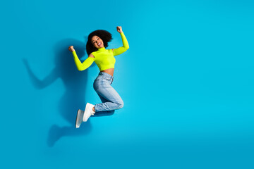 Joyful young woman jumping with excitement against a vibrant blue background, wearing bright neon...