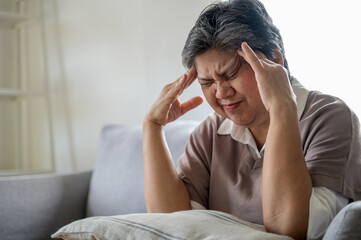 An elderly Asian woman is sitting on a sofa at home, experiencing a headache, dizziness, and a...