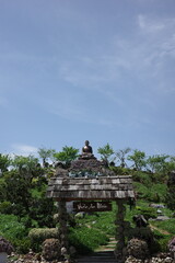 Naklejka premium A serene Buddha statue sits atop a landscaped hill above a rustic wooden gate in a peaceful Zen garden, surrounded by lush greenery and clear blue sky. A perfect symbol of harmony, nature