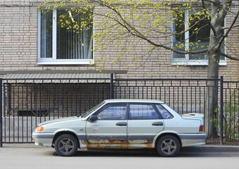 An old rusty silver car stands near the lattice fence of a brick housem, Dybenko Street, St....