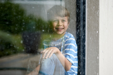 Happy, child and watching rain by window for sensory experience, imagination and winter weather view. Boy kid, smile and thinking in home with daydreaming, memory and checking scenery with reflection